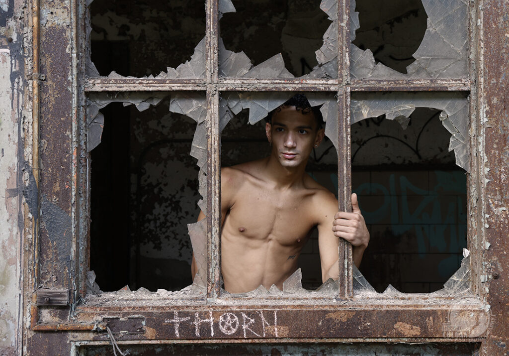 shirtless man poses behind broken windows of a metal door