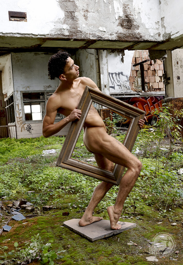 naked man with empty picture frame between his legs in old building