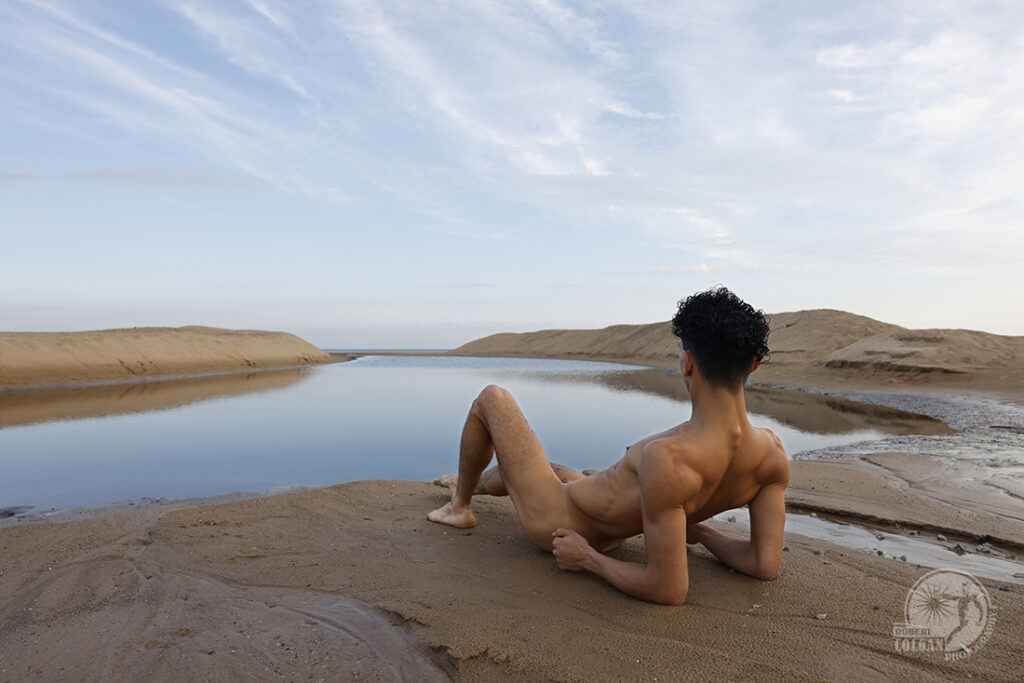 nude man reclining on a beach overlooking a calm inlet of water