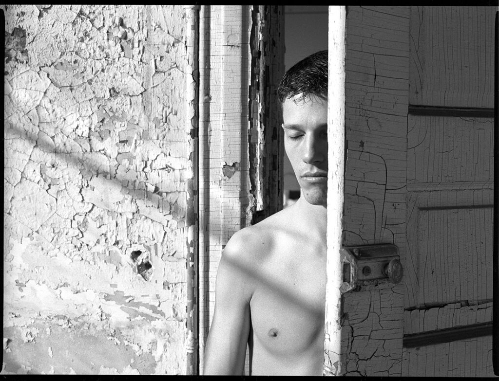 shirtless man stands with eyes closed in doorway of old building