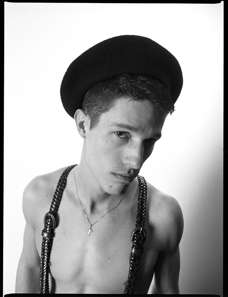 shirtless man wearing suspenders looks sidewise at camera