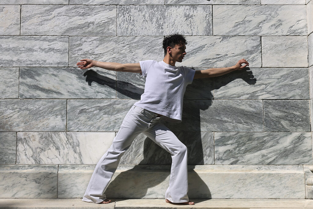 man dressing in white stretches in dance pose beside white marble wall