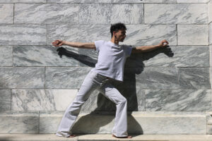 man dressing in white stretches in dance pose beside white marble wall