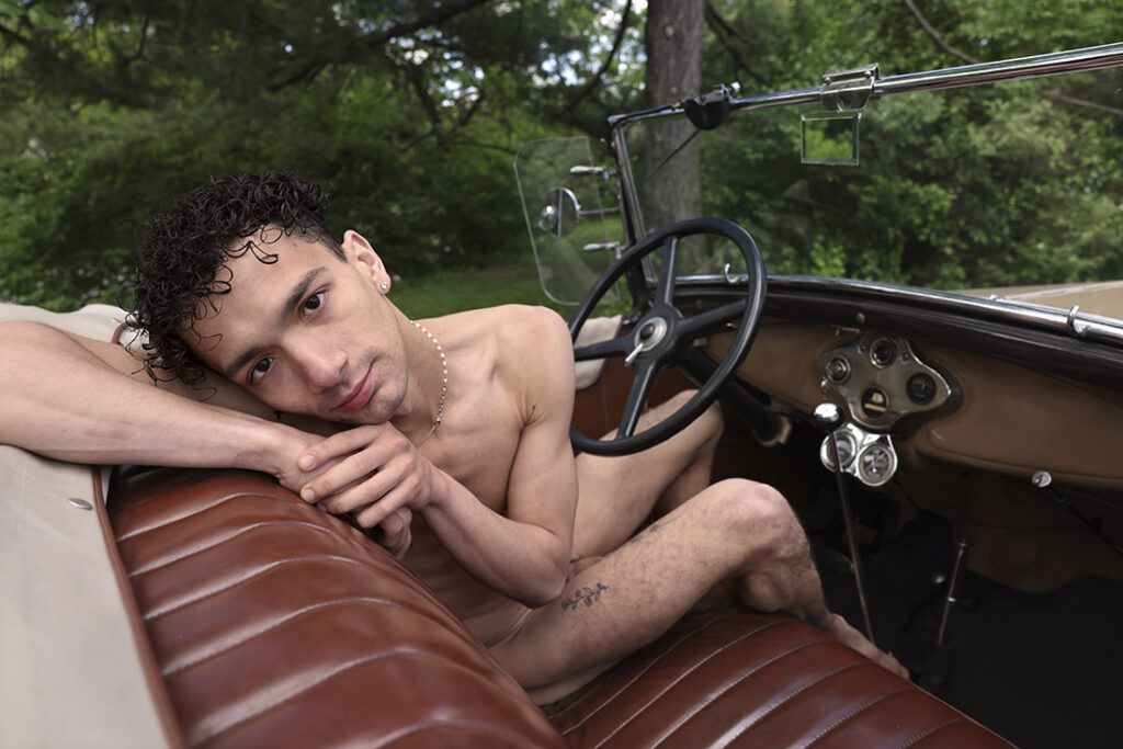 nude man leans across leather seat of vintage car to stare into the camera