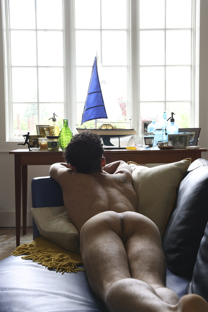 nude man outstretched on sofa looking out a huge window with miscellaneous items on a table in front of it.