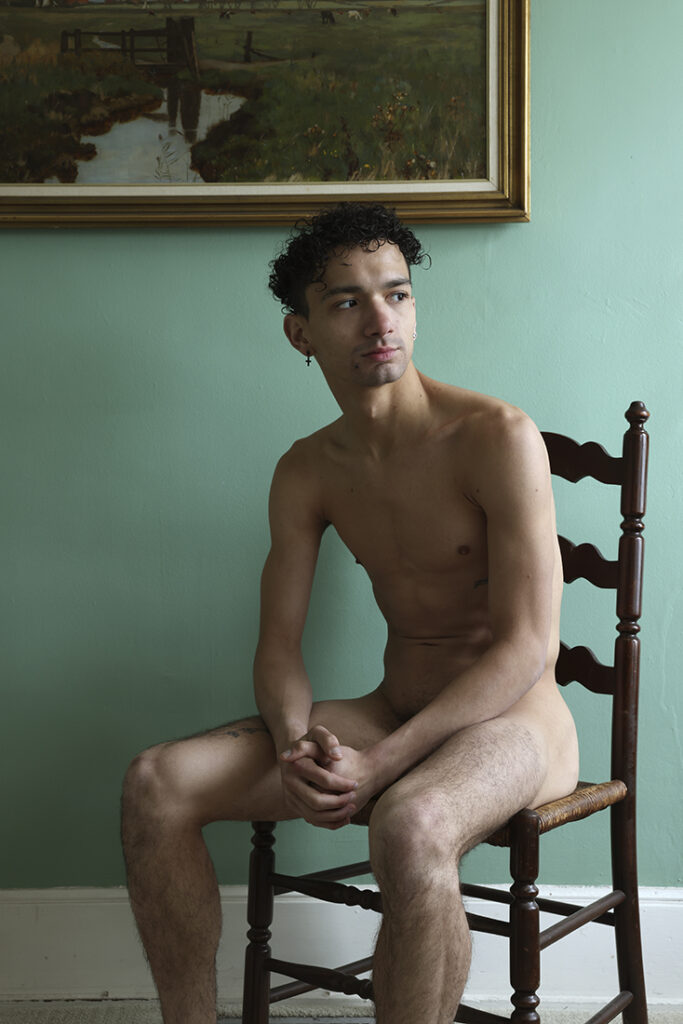 nude man sits in ladderback chair in green room