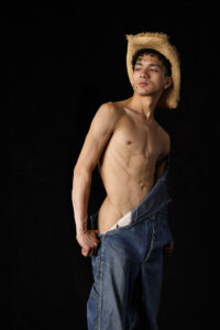 studio shot of man in overalls with straw hat seductively exposing his naked hip