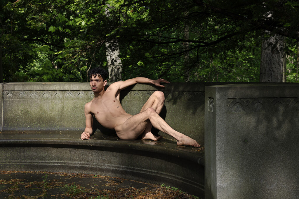 nude man lounges on classic stone bench with a wooded scene behind him
