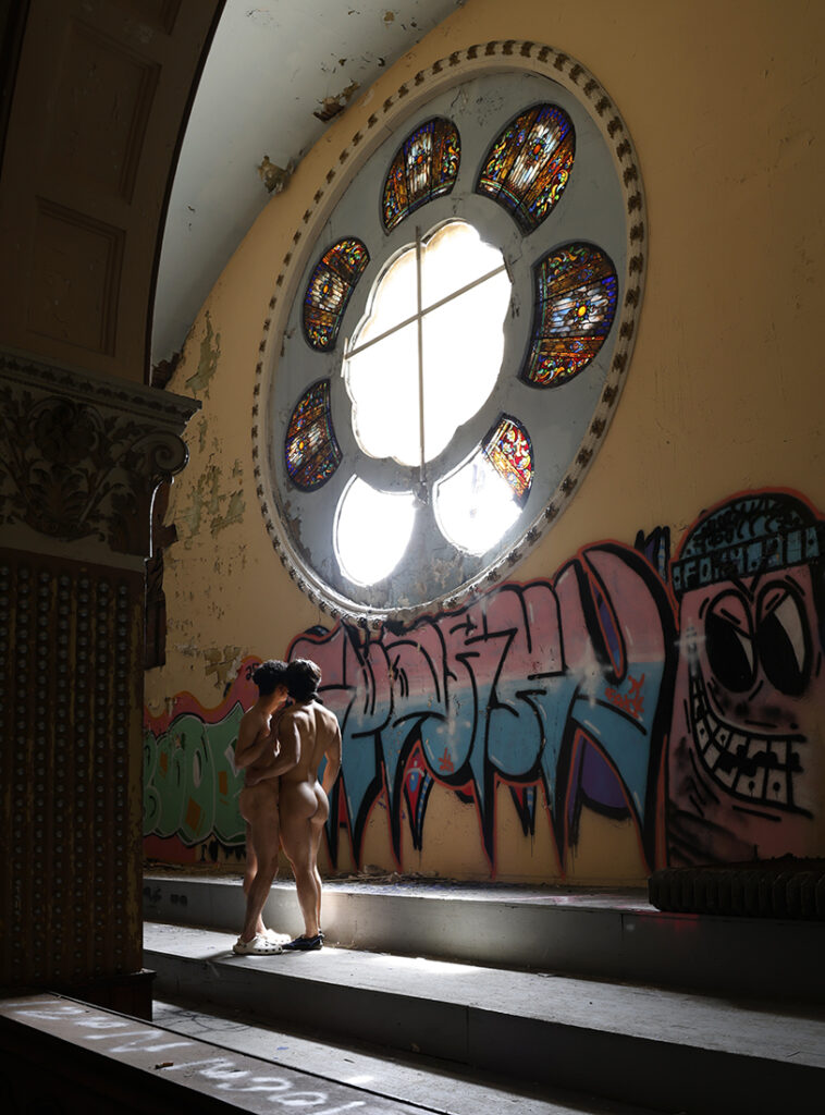 two nude men stand facing a huge stained glass rose window in abandoned church
