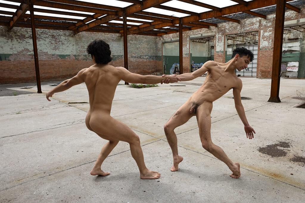two nude men grasping hands and extending their bodies in dance while in abandoned building