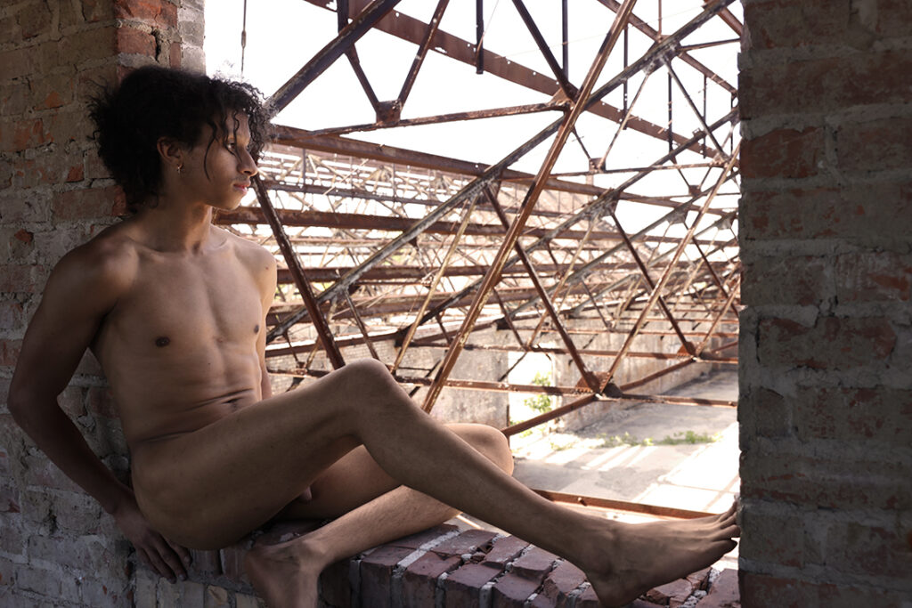 nude man sits in window opening overlooking lots of girders