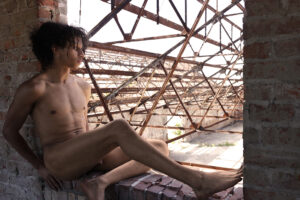 nude man sits in window opening overlooking lots of girders