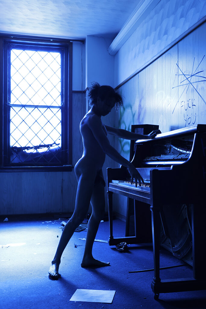 nude man pplays piano while bathed in blue stained glass light, captured from the side