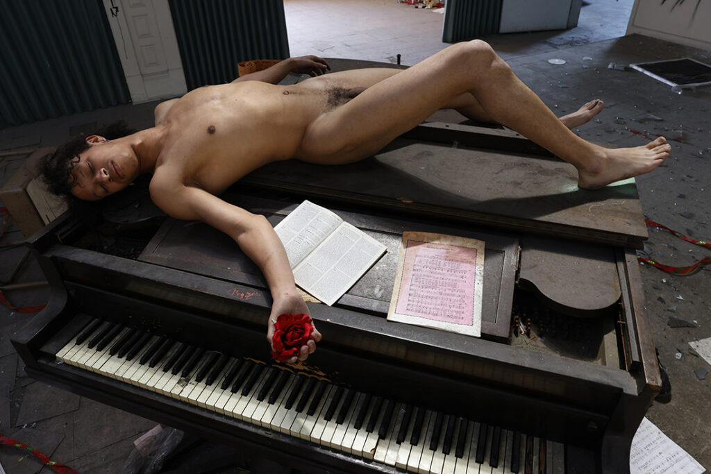Nude man lays on top of piano while clutching a red rose