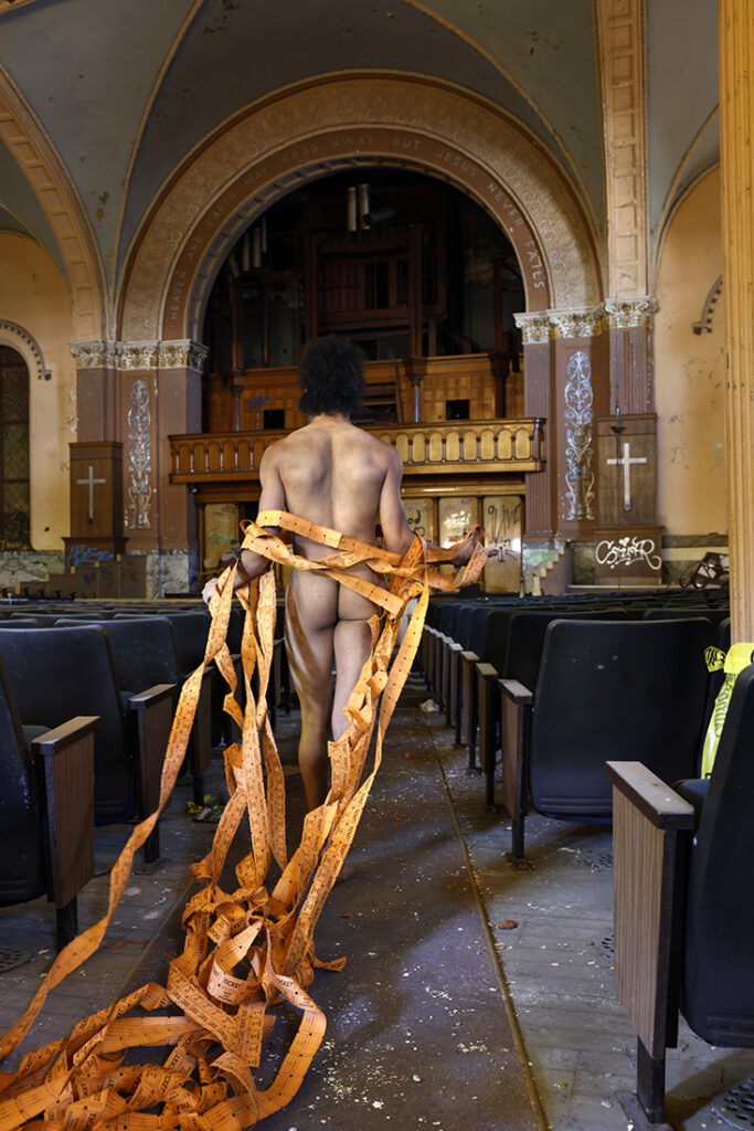 nude man tethered with streamers of paper tickets walks of aisle of abandoned church