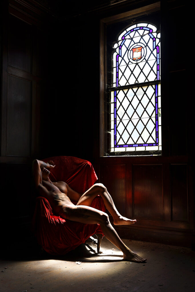 Darkened image of naked man sitting in red chair with light from a stained glass window