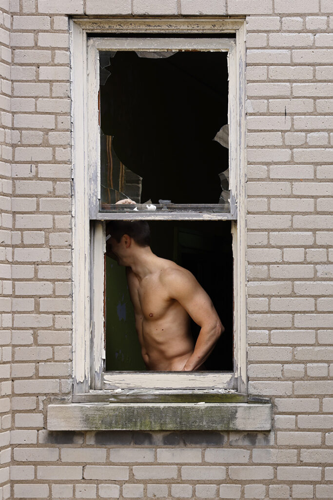 nude man in broken window obscures his face from the viewer